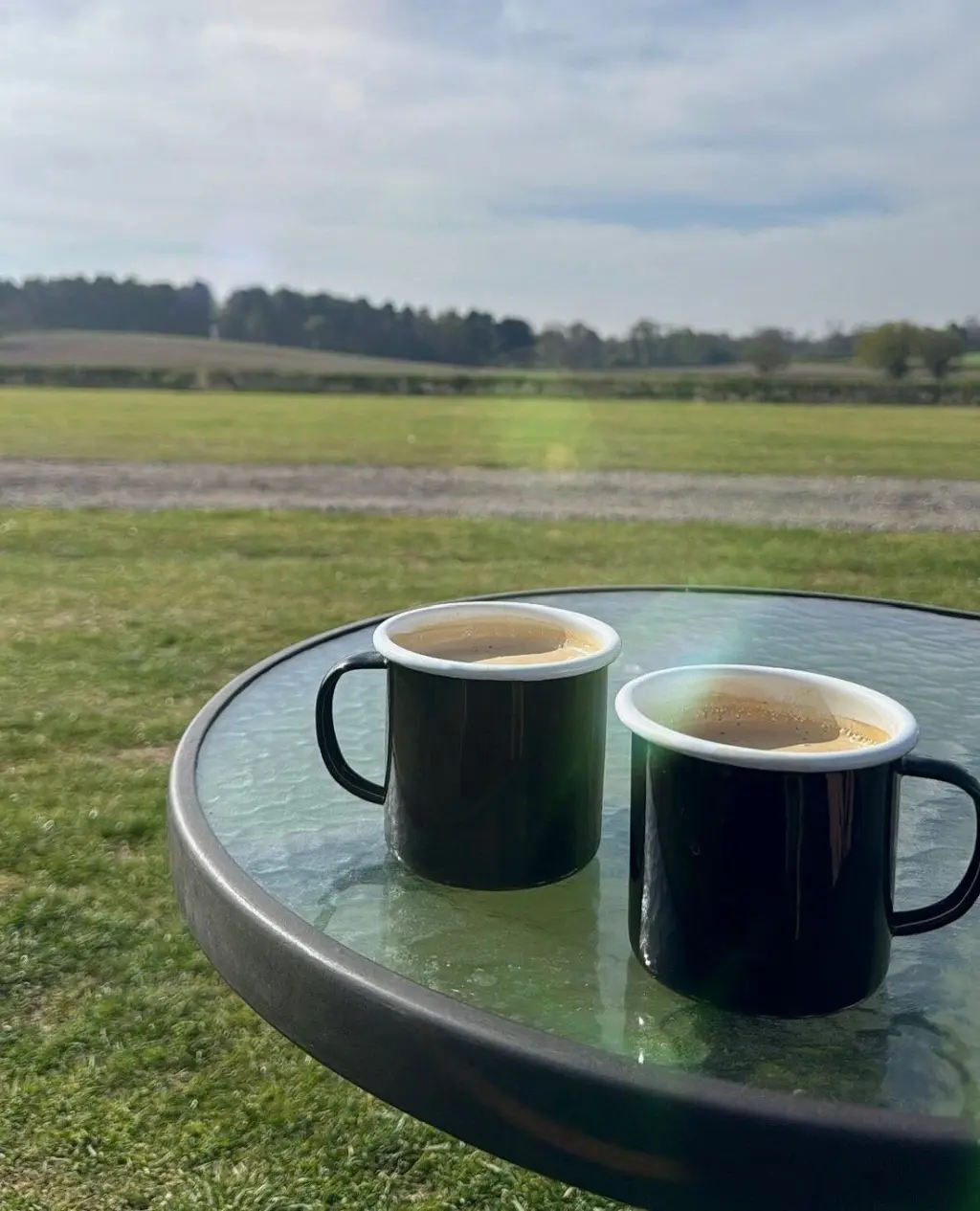 Two black enamel mugs of coffee on a glass table in a field with trees in the background.