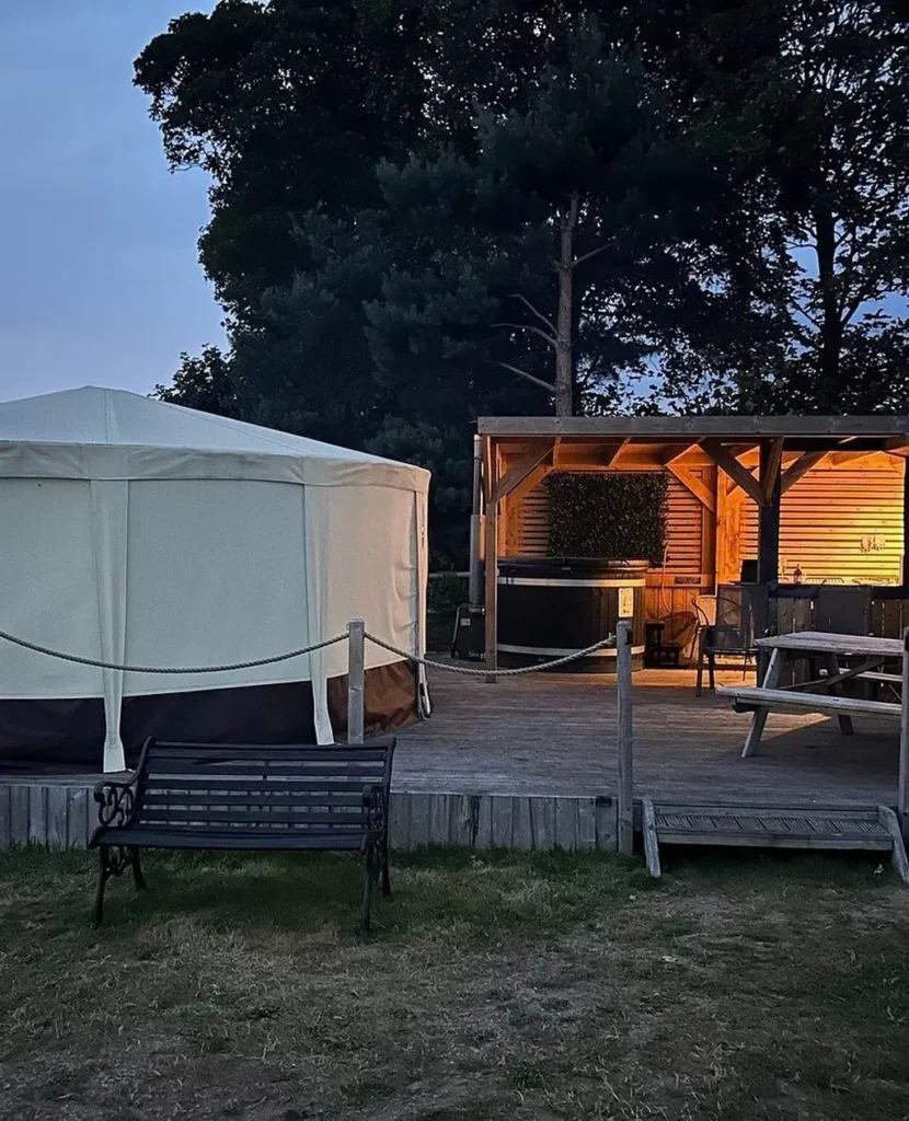 A yurt and a wooden deck with a hot tub, picnic bench, and lit shelter at dusk.